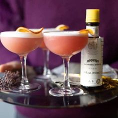 two martinis with orange peel garnish sit on a tray next to a bottle of booze