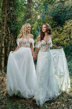 two women in wedding gowns standing next to each other holding hands and looking at each other