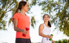 two women are smiling while holding dumbs and looking at each other with trees in the background