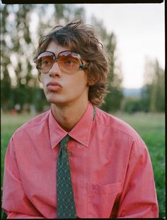 a man wearing glasses and a tie sitting in the middle of a field with trees behind him