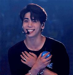 a young man with black hair and piercings smiles while holding his hands together in front of him