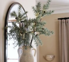 a vase filled with pine branches on top of a table next to a mirror and window