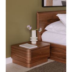 a small white vase with flowers on top of a nightstand next to a large bed