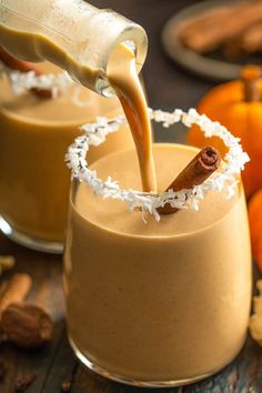 two glasses filled with pumpkin spiced smoothie on top of a wooden table next to oranges and cinnamon sticks