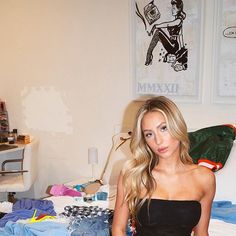 a beautiful young blond woman sitting on top of a bed next to a pile of clothes