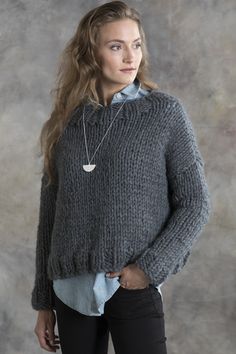 a woman standing in front of a gray wall wearing a sweater and black pants with her hands on her hips