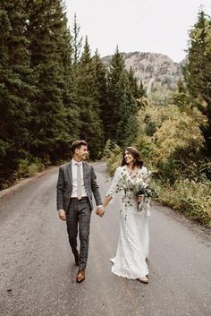 a bride and groom holding hands walking down the road in front of some pine trees