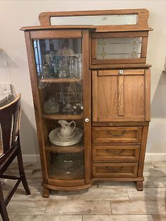 a wooden china cabinet with glass doors and drawers on the bottom, next to a chair