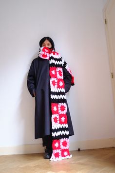 a woman standing in front of a white wall wearing a black coat and red scarf