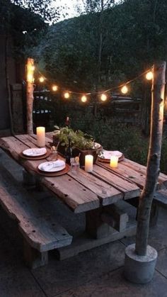 a wooden table with candles and plates on it in front of some lights hanging from the trees