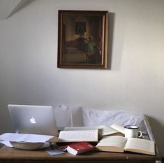 an open book on a table next to a laptop and coffee cup with a painting above it