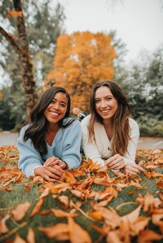 two women laying on the ground with leaves in front of them and smiling at the camera