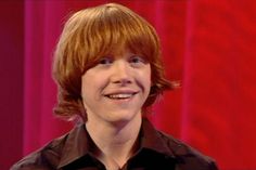 a young man with red hair smiles at the camera while standing in front of a red curtain