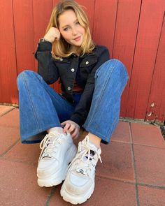 a woman sitting on the ground with her feet up and wearing white tennis shoes in front of a red wall