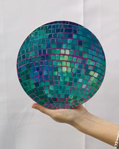 a hand holding up a green and purple disco ball with squares on the top, in front of a white background