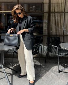 a woman in black blazer and white pants holding a black handbag while standing next to a table