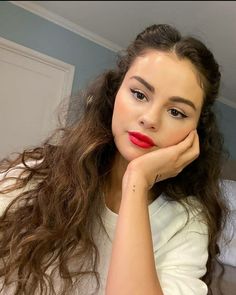 a woman with long hair and red lipstick looks at the camera while holding her hand on her chin