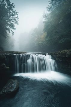 a waterfall in the middle of a forest filled with lots of trees and foggy skies