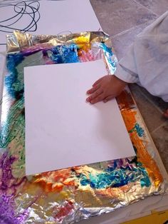 a person holding a piece of paper on top of tin foil with paint all over it