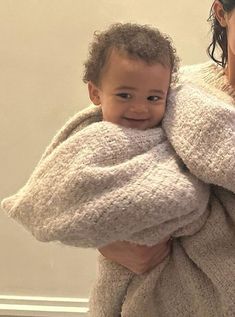 a woman is holding a baby wrapped in a blanket and smiling at the camera while standing next to a wall