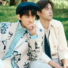 two young men sitting on the grass in front of trees and one is wearing a hat