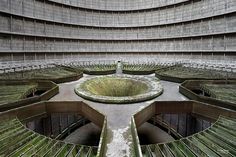 a circular building with wooden slats in the center and concrete walls around it, surrounded by grass