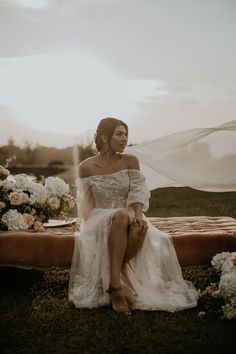 a woman sitting on top of a bed in a field next to flowers and a veil