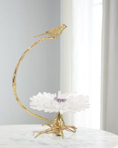 a white and gold flower with a bird on it sitting on a table next to a window