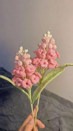 a hand holding a pink flower in front of a white wall and gray bedding