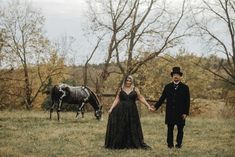 Non-traditional alternative bride holding hands with her groom in a vintage Victorian inspired shoot. they are posed outside in a field with a horse behind them who has a painted skeleton on the coat. Spooky Halloween Wedding, Bride In Black, Dapper Grooms