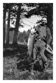 black and white photograph of a man holding a fish in his hand while standing next to trees