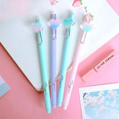 three pastel pens sitting on top of a pink surface next to an open notebook