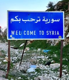 a welcome to syria sign sitting on the side of a road in front of a rocky hill