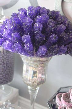 purple flowers in a silver vase next to cookies on a table with other desserts