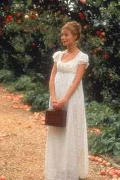 a woman in a white dress holding a brown purse standing next to an apple tree