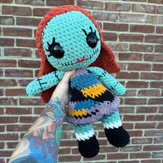 a hand holding a crocheted doll in front of a brick wall