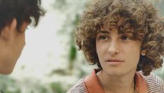 a young man with curly hair is talking to another person in a striped shirt and vest