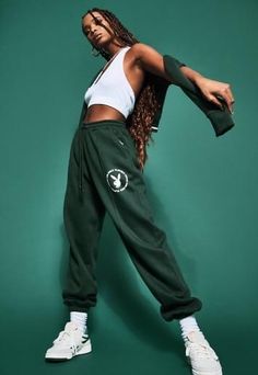 a woman with long hair wearing green sweatpants and white sneakers, leaning against a green wall