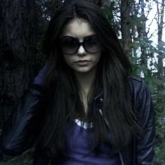 a woman wearing sunglasses and a leather jacket standing in the woods with her hand on her head