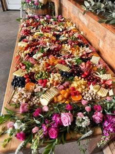 an assortment of fruits and cheeses on a long wooden table with greenery in the background