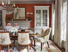 an elegant dining room with red walls and white curtains on the windowsills is furnished with floral upholstered chairs