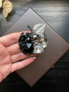 a hand holding an apple brooch with leaves and jewels on it's side