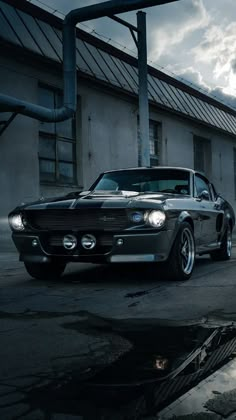 an old black car parked in front of a building with cloudy skies behind it and water puddles on the ground