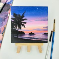 a painting on an easel next to paintbrushes and watercolor paper with a palm tree in the foreground