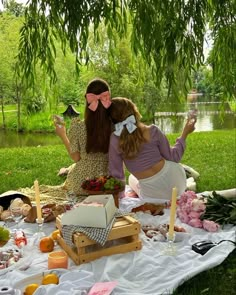 two women sitting on a blanket in front of a lake with food and drinks around them