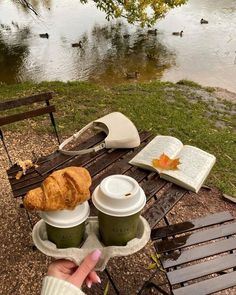 there is a croissant, coffee and book on the picnic table by the river