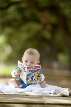 a baby is sitting on a blanket reading a comic book while looking at the camera