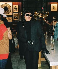 a man in a black suit and white hat standing next to a counter with wine glasses on it