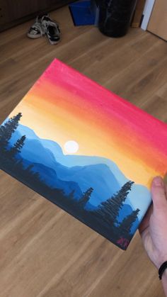 a person holding up a piece of art with mountains in the background and trees painted on it