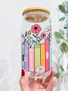 a hand holding a glass jar with books and flowers painted on it